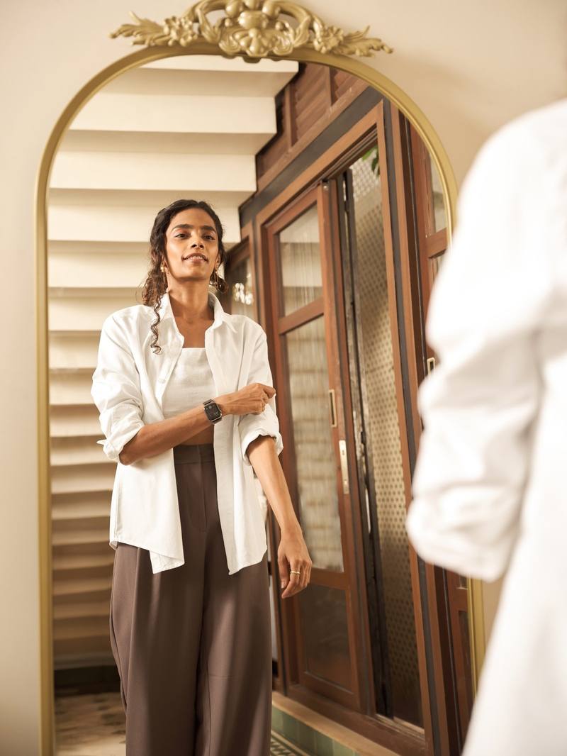 A woman in a white shirt and brown trousers by an arched mirror